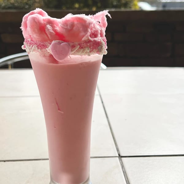 Pink milkshake topped with cotton candy, sprinkles, and a heart candy on a tiled table.