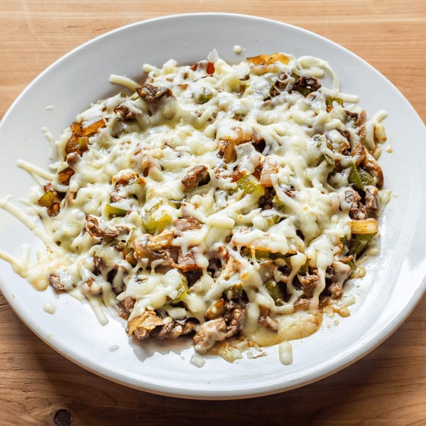 A white plate holds a Mexican dish with melted cheese, meat, green peppers, and onions.