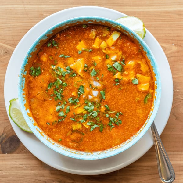 Overhead view of a vibrant orange Mexican soup in a blue bowl, garnished with cilantro and lime.