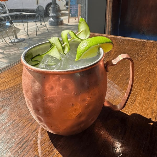 A copper mug filled with a cocktail, ice, cucumber ribbons, and a lime wedge on a wooden table.