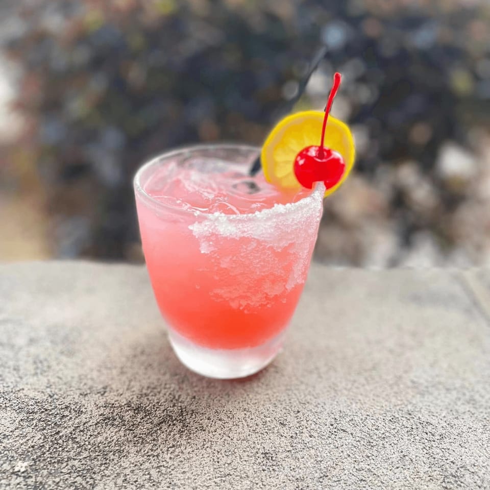 Pink margarita with a salted rim, lemon slice, and cherry, on a textured surface.