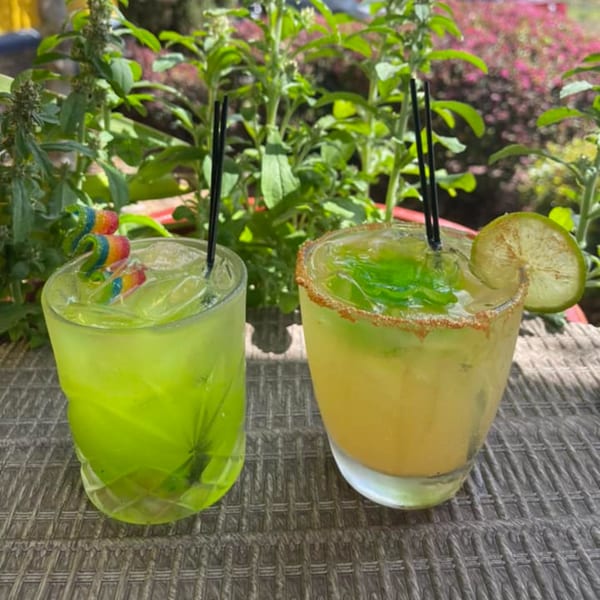 Two vibrant Coyoacan Mexican Restaurant cocktails, one green with candy, one yellow with a lime, on a patio.