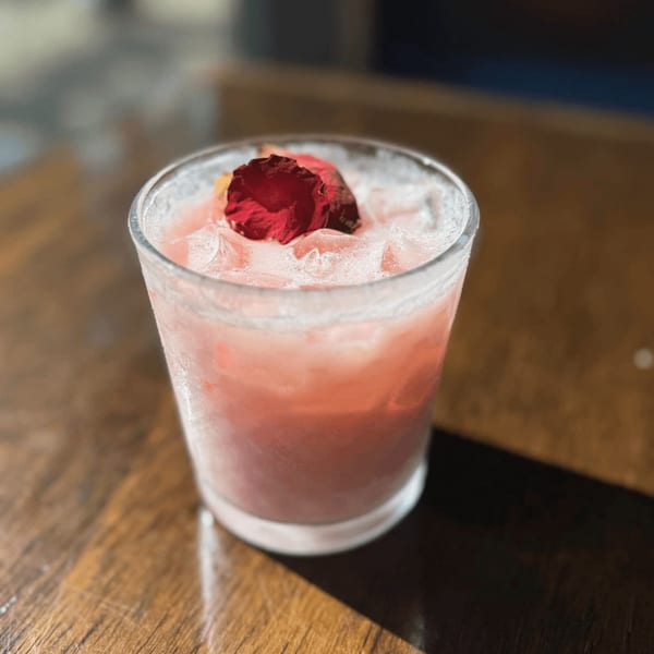 Pink cocktail with ice and a red rose petal garnish on a wooden table.