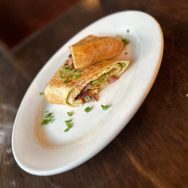 Two Mexican breakfast burritos, one cut open to show eggs, bacon, and avocado, on a white plate.
