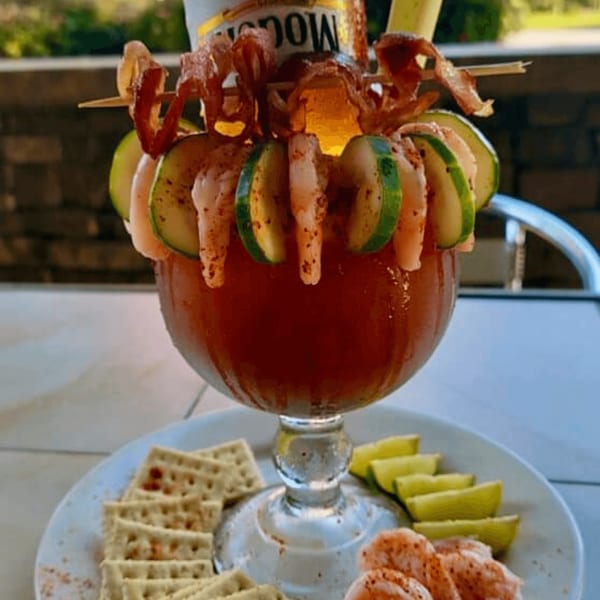 A large Michelada cocktail garnished with shrimp, cucumber, bacon, and a beer bottle, served with crackers and lime.