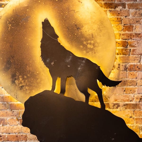 A metal cutout of a howling wolf against a glowing, textured full moon on a brick wall.