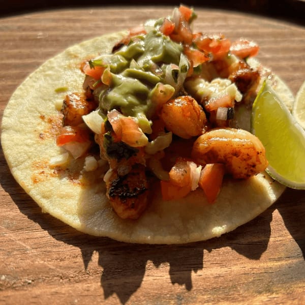 A shrimp taco with guacamole, pico de gallo, and a lime wedge on a wooden surface.