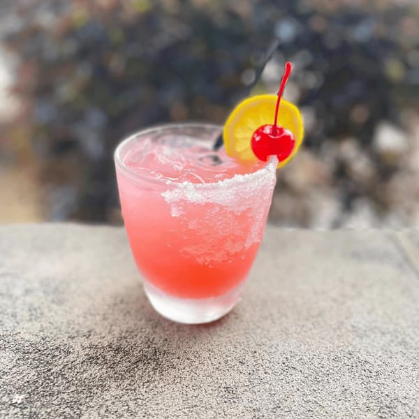 Pink margarita with a salted rim, lemon slice, and cherry, on a textured surface.